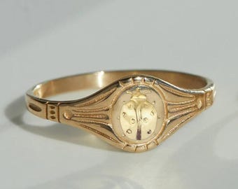 Può includere: Anello color oro con un design dettagliato di coccinella al centro. L'anello ha una faccia ovale con una coccinella in rilievo e linee decorative. La fascia è in oro lucido semplice.