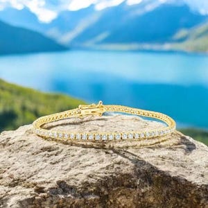 May include: A gold tennis bracelet with numerous small, clear gemstones. The bracelet is displayed on a rock with a scenic background of a lake and mountains.