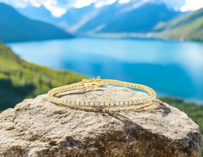 May include: A gold tennis bracelet with numerous small, clear gemstones. The bracelet is displayed on a rock with a scenic background of a lake and mountains.