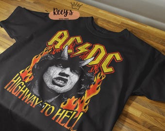 May include: Black t-shirt featuring the band AC/DC with a graphic of a man with horns and flames. The text "AC/DC" is in yellow, and "HIGHWAY TO HELL" is in orange. The shirt is on a wooden surface.