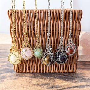 Può includere: Una collezione di collane con pietre preziose di vari colori, tra cui giallo, rosa, verde e marrone, esposte in un cesto di vimini. Ogni collana presenta una catena e una gabbia metallica decorativa.