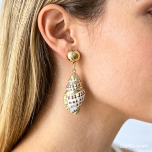 May include: Gold-colored stud earrings with a dangling seashell design. The seashells are white with brown markings and gold accents. The earrings are a unique and elegant accessory.