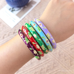 May include: A set of five stackable bangles in purple, red, green, light blue, and lavender. Each bangle features a glossy enamel finish with gold-tone floral patterns, including small leaves and blossoms distributed across the outer surface.