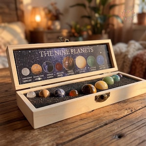 May include: A wooden box displaying a collection of gemstone spheres representing the nine planets. The box features a printed illustration of the planets with their names. The gemstones are arranged on a black foam insert. The box is open, revealing the gemstones and the planet illustration.