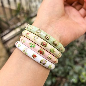 May include: Four colorful bangles with floral designs. The bangles are in shades of white, pink, cream, and green, with gold-colored accents. The floral patterns include red, yellow, and green details. The bangles are approximately 0.5 inches wide.