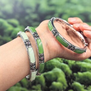 May include: Three silver bangles with jade stones. The bangles have a floral design with small red and green accents. The jade stones are light green and white. The bangles are on a wrist.