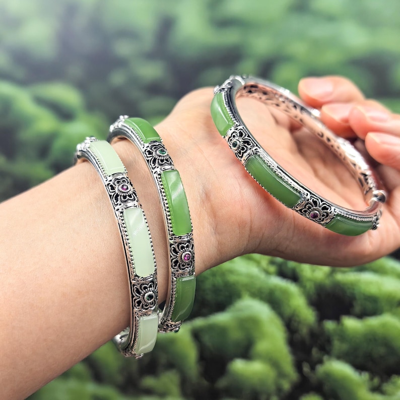 May include: Three silver bangles with jade stones. The bangles have a floral design with small red and green accents. The jade stones are light green and white. The bangles are on a wrist.