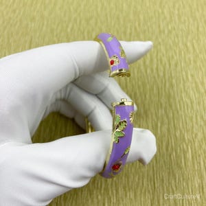 May include: A hinged bangle bracelet featuring a lavender enamel finish with gold-tone metal edges. The surface is decorated with small floral motifs in red and green enamel. The bracelet is shown in an open position, revealing the gold-tone hinge.