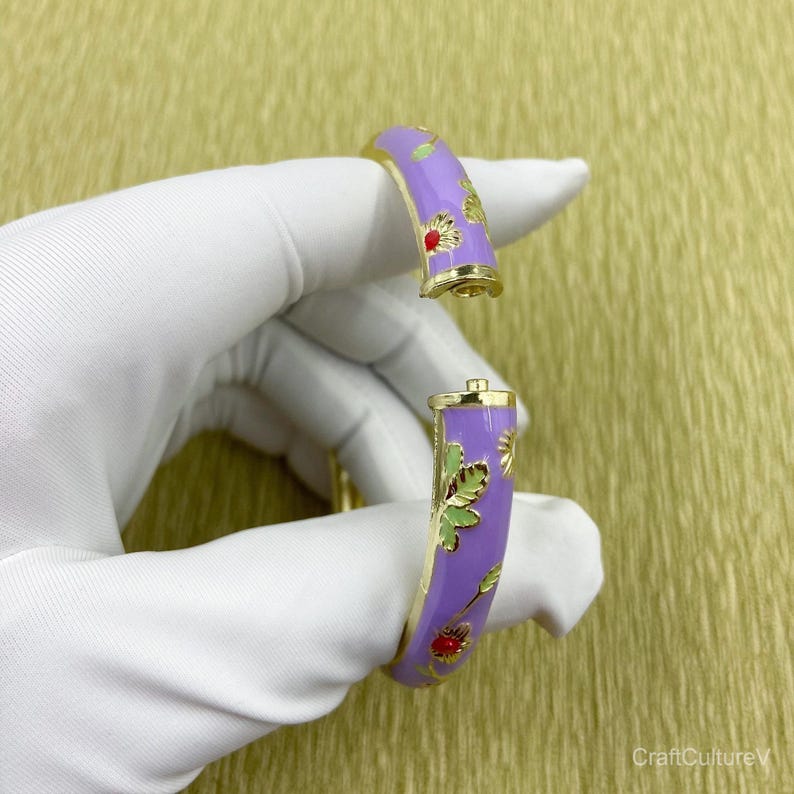 May include: A hinged bangle bracelet featuring a lavender enamel finish with gold-tone metal edges. The surface is decorated with small floral motifs in red and green enamel. The bracelet is shown in an open position, revealing the gold-tone hinge.