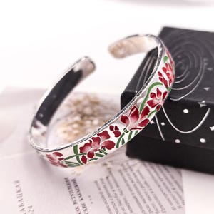 May include: A silver cuff bracelet with a floral design. The bracelet features red flowers with green stems and leaves, set against a white background. The bracelet is displayed on a printed page and a black box.