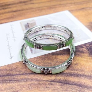 May include: Two silver bangles with green jade stones and intricate floral designs. The bangles have a rectangular jade stone in the center and small red stones. The background is a wooden surface with a white paper.