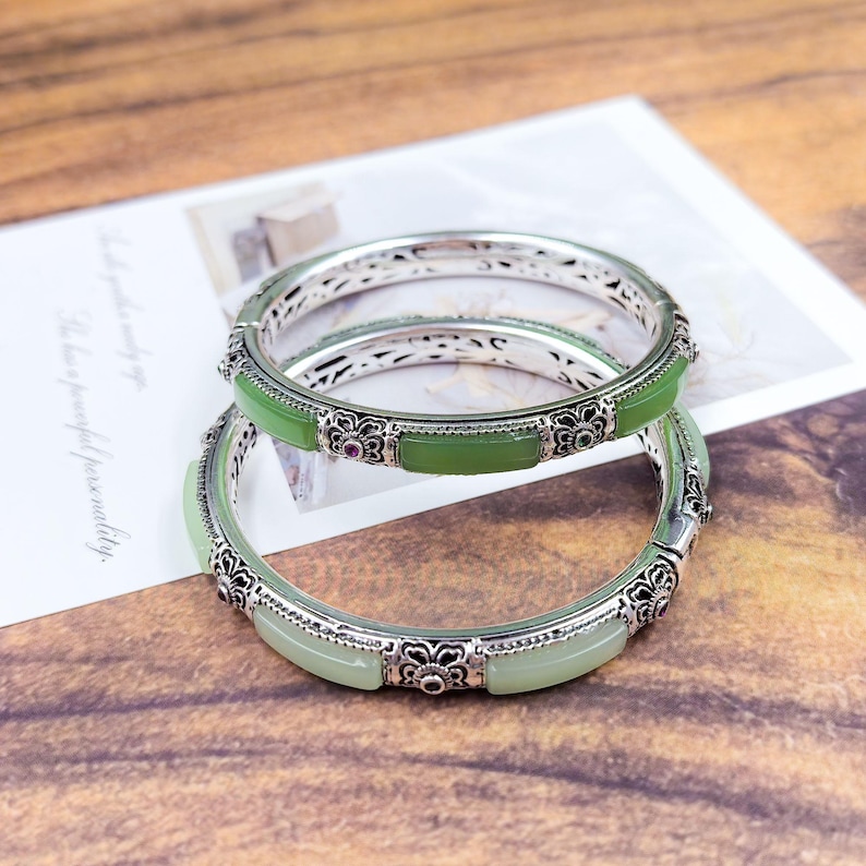 May include: Two silver bangles with green jade stones and intricate floral designs. The bangles have a rectangular jade stone in the center and small red stones. The background is a wooden surface with a white paper.