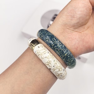 Puede incluir: Dos pulseras decorativas. Una es blanca con diseños de mariposas doradas y detalles dorados. La otra es de color verde azulado oscuro con diseños de mariposas doradas. Las pulseras se llevan en la muñeca.