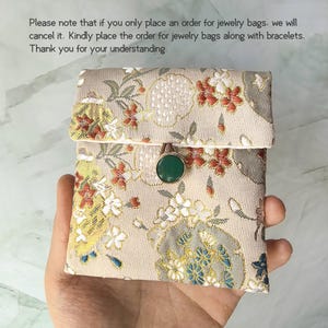 May include: A small square fabric pouch with a beige base featuring a woven floral pattern in red, white, and blue. The pouch has a flap closure secured by a round green button. Text above reads: Please note that if you only place an order for jewelry bags, we will cancel it. Kindly place the order for jewelry bags along with bracelets. Thank you for your understanding.