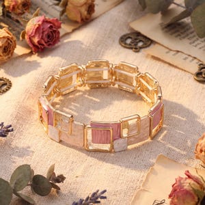 May include: A gold-tone stretch bracelet featuring a series of connected square links. The links alternate between solid gold-tone metal, soft pink enamel, and cream-colored enamel, all with a textured finish. The bracelet has a flexible design.