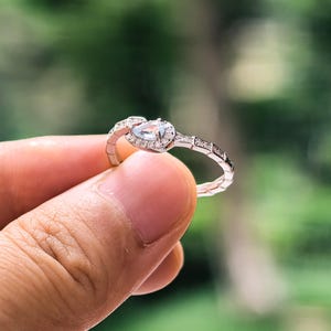 Puede incluir: Un anillo plateado con una piedra preciosa ovalada de color azul claro en el centro, rodeada de pequeñas piedras transparentes. La banda presenta un diseño texturizado con piedras pequeñas adicionales. El anillo se sostiene contra un fondo verde borroso.