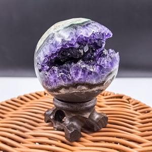 May include: A spherical amethyst geode, showcasing a deep purple crystal interior. The geode is mounted on a dark brown wooden stand, set against a neutral background. The geode is approximately 4 inches in diameter.