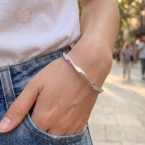 May include: A polished silver-tone bangle bracelet featuring a continuous, fluid, and undulating wave-like design. The metal surface is smooth and reflective, creating a sleek, curved silhouette that wraps around the wrist with a subtle, organic form.