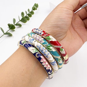 Op de afbeelding: Een verzameling kleurrijke emaille armbanden met bloemmotieven. De armbanden hebben verschillende kleuren, waaronder rood, blauw, wit en turquoise, met goudkleurige accenten en strass-steentjes. Elke armband heeft een diameter van ongeveer 6,35 cm.