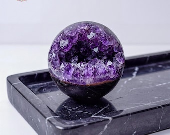 May include: A spherical amethyst geode, displaying a rich purple crystal interior. The geode sits on a dark, rectangular marble tray. The geode's exterior is a dark colour, contrasting with the bright purple crystals inside.