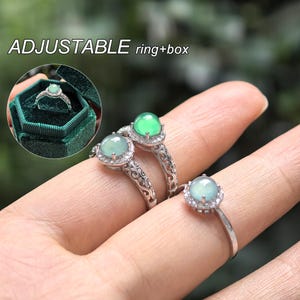 Op de afbeelding: Zilveren ringen met een ronde, lichtgroene edelsteen in het midden en een halo van kleinere, heldere stenen. De ringen worden op een hand getoond, met één ring in een groene fluwelen ringdoos. De tekst "ADJUSTABLE ring+box" is zichtbaar.