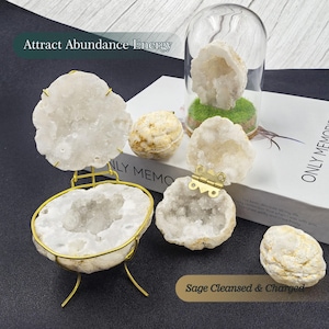 Könnte beinhalten: Eine Sammlung weißer und beiger Geoden, einige davon auf goldfarbenen Ständern und in einer Glaskuppel präsentiert. Der Text "Attract Abundance Energy" und "Sage Cleansed & Charged" ist sichtbar. Die Geoden variieren in Größe und Form.