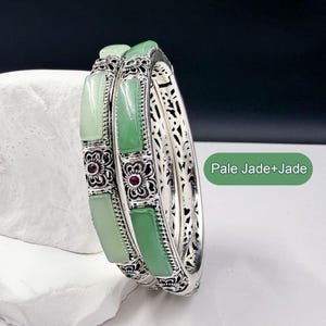 Puede incluir: Dos brazaletes de plata con incrustaciones de jade verde pálido y detalles florales. Los brazaletes tienen un diseño de filigrana en los laterales y pequeñas gemas rojas. El texto "Pale Jade+Jade" está en la imagen.