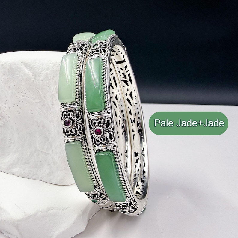 May include: Two silver bangles with pale green jade inserts and floral accents. The bangles have a filigree design on the sides and small red gemstones in the center of the floral designs. The text "Pale Jade+Jade" is in the image.