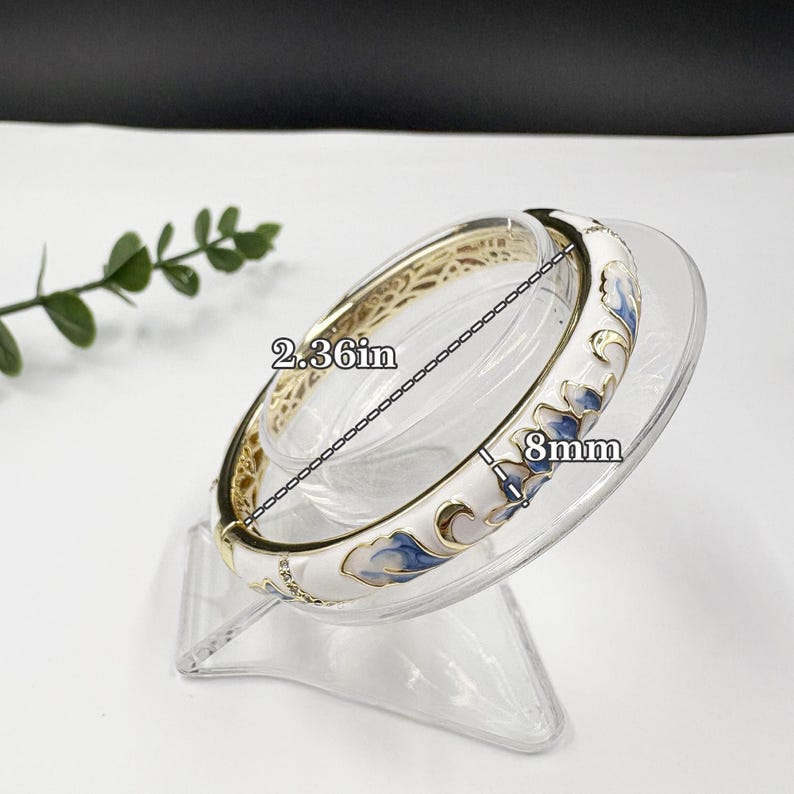 May include: A hinged bangle bracelet featuring a white enamel surface with blue floral patterns and gold-tone accents. The bracelet has a diameter of 2.36 inches and a width of 8mm, as indicated by the text labels on the image.