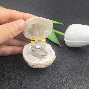 Könnte beinhalten: Eine weiße Geoden-Ringbox mit einem goldenen Scharnier, geöffnet, um einen silbernen Verlobungsring mit einem großen Mittelstück und einem kleineren Band zu enthüllen.