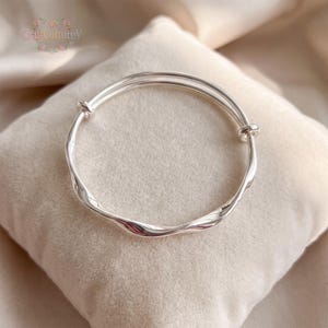 May include: A polished silver-tone bangle bracelet featuring a continuous twisted, wavy design. The circular metal band includes two small sliding knots that allow for size adjustment. The surface is smooth and reflective, catching light.