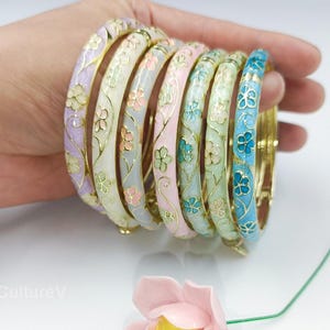 May include: A collection of colorful enamel bangles in shades of lavender, white, pink, green, and blue. Each bangle features a floral design with gold-colored accents. The bangles are held in a hand, showcasing their intricate details and vibrant colors.