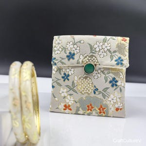 May include: Two cream-colored bangles with gold floral designs are next to a small, floral-patterned pouch. The pouch has a green gemstone button closure. The pouch's fabric features white, blue, and orange flowers on a light green background.