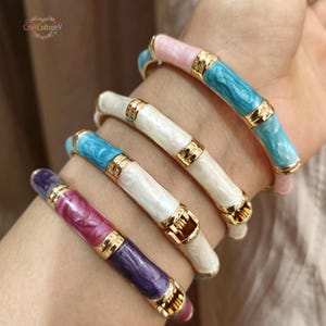 May include: A collection of colorful enamel bangles with gold-tone accents. The bracelets feature shades of pink, blue, white, and purple. The bracelets are worn on a wrist, showcasing the jewelry's design and color variations. The bracelets have a bamboo-like design.