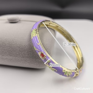 May include: A gold-tone hinged bangle bracelet featuring a lavender enamel finish with a floral pattern in green and red. The bracelet measures 2.36 inches in diameter and has a band width of 8mm, as indicated by the visible measurement markings.