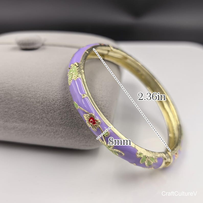 May include: A gold-tone hinged bangle bracelet featuring a lavender enamel finish with a floral pattern in green and red. The bracelet measures 2.36 inches in diameter and has a band width of 8mm, as indicated by the visible measurement markings.