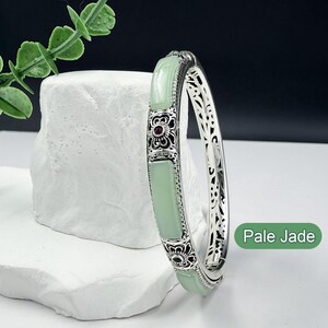 May include: A silver bangle bracelet with pale jade panels and intricate floral designs. The bracelet features a rectangular jade stone and a small red gem in the center of the floral design. The text "Pale Jade" is visible.