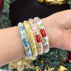 Pode incluir: Quatro pulseiras florais esmaltadas em tons de azul, amarelo, rosa e vermelho. Cada pulseira apresenta um design floral diferente com detalhes dourados sobre um fundo branco. As pulseiras são usadas no pulso.