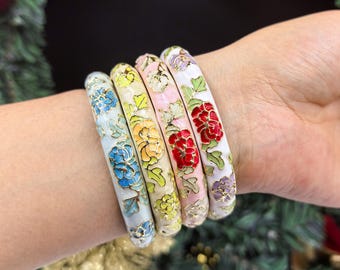 Könnte beinhalten: Vier Emaille-Armreifen mit Blumenmustern in Blau, Gelb, Rosa und Rot. Jeder Armreif hat ein anderes Blumendesign mit goldenen Akzenten auf weißem Hintergrund. Die Armreifen werden am Handgelenk getragen.