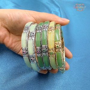May include: A collection of jade bangles in varying shades of green and yellow. The bangles have ornate silver-colored metalwork with floral and gemstone accents. The jade pieces are rectangular and set within the metal frames. The bangles are stacked together.