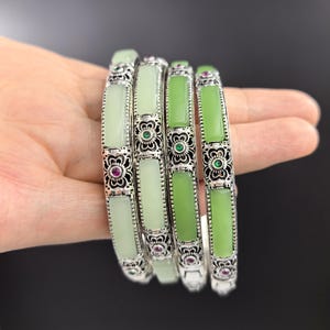 May include: Four silver bangles with rectangular jade stones in varying shades of green. The silver bands feature intricate floral designs with small red and green gemstones. The bangles are stacked together.