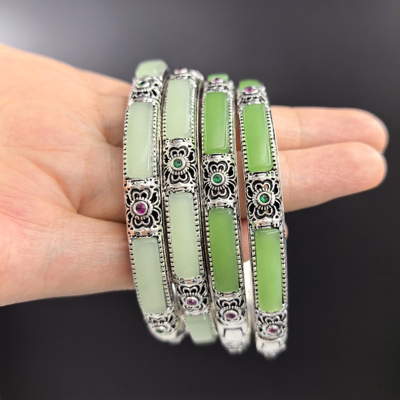 May include: Four silver bangles with rectangular jade stones in varying shades of green. The silver bands feature intricate floral designs with small red and green gemstones. The bangles are stacked together.