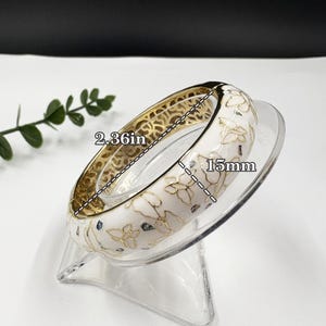 May include: A hinged bangle bracelet featuring a white enamel surface with gold-toned butterfly outlines and scattered dark flecks. The bracelet has a diameter of 2.36 inches and a width of 15mm, with an intricate gold-toned filigree pattern on the interior.
