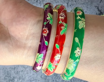 May include: Three colorful bangles with floral designs. The bangles are green, red, and purple with gold accents. The bangles are stacked on a person's wrist.