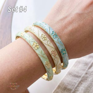 Può includere: Set di quattro braccialetti smaltati in colori pastello. Ogni braccialetto presenta un motivo floreale con dettagli dorati. I braccialetti sono in verde chiaro, crema e blu, con dettagli floreali rosa e turchese. Il testo "Set #4" è visibile.