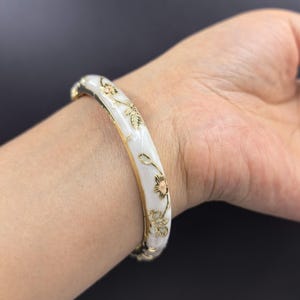 May include: A hinged bangle bracelet featuring a smooth white enamel surface decorated with delicate gold-tone floral engravings. The metal edges of the bracelet are finished in a polished gold tone, providing a contrast to the white enamel inlay.