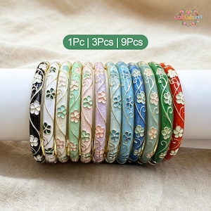 Könnte beinhalten: Eine Sammlung von neun Emaille-Armreifen in verschiedenen Farben, darunter Schwarz, Weiß, Grün, Lila, Blau und Rot. Jeder Armreif ist mit einem floralen Design und goldfarbenen Akzenten verziert. Der Text "1Pc | 3Pcs | 9Pcs" ist zu sehen.