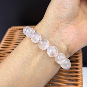Könnte beinhalten: Ein Armband aus klarem Quarz mit sechs runden Perlen. Die Perlen sind auf einem rosa elastischen Band aufgefädelt. Das Armband wird am Handgelenk getragen. Die Quarzperlen haben einen leicht rosa Farbton und innere Adern.
