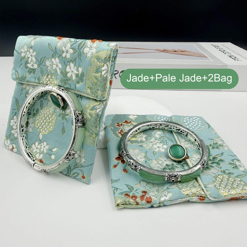 May include: Two jade bangles with silver filigree detailing and a matching jade cabochon are displayed on a floral patterned pouch. The pouches are a light blue-green color. The text "Jade+Pale Jade+2Bag" is visible.