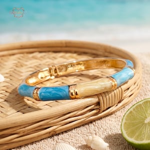 Op de afbeelding: Gouden scharnierende armband met blauwe en crèmekleurige emaille accenten. De armband heeft een bamboe-stijl ontwerp met goudkleurige metalen accenten. De armband wordt gepresenteerd op een geweven dienblad met een strandachtergrond.